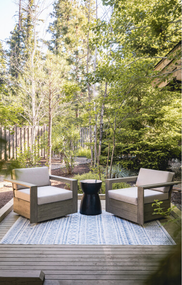 Two wooden armchairs with cushions face each other on a patterned rug in the sunny, tree-lined outdoor deck area of Suncadia Resort. A small black table sits between them, offering a perfect spot to enjoy the serene atmosphere.