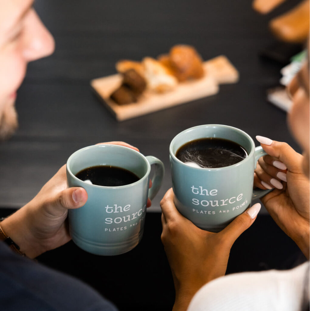 Two people holding green mugs with "the source" printed on them, filled with black coffee. A wooden board with pastries is visible in the background, capturing a cozy moment reminiscent of mornings in Suncadia.