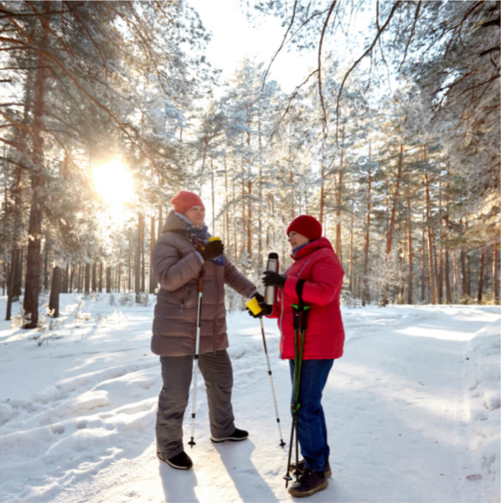 Two people dressed in winter clothing stand on a snowy path in a sunlit forest at Suncadia Resort Washington, holding walking poles.