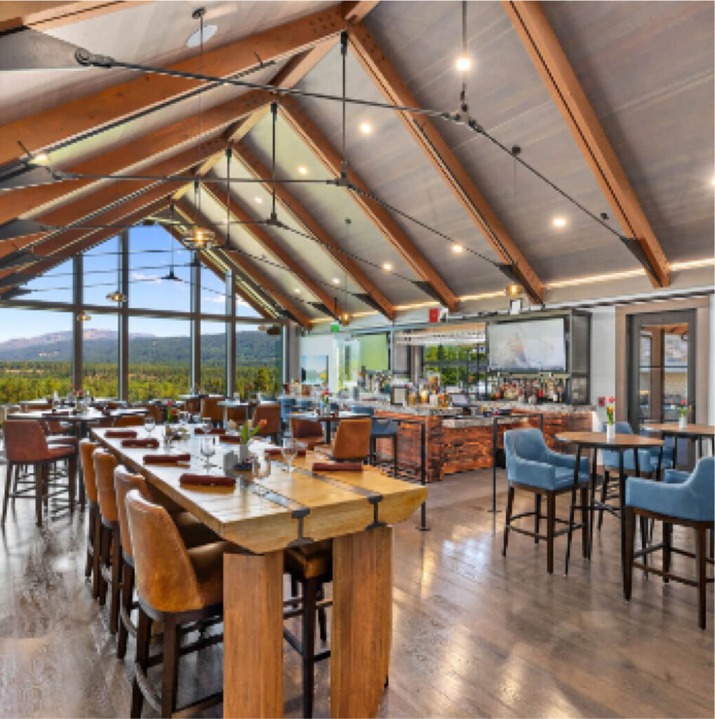 A modern restaurant interior at Suncadia Resort features wooden beams, tables elegantly set for dining, and high chairs at a bar. Large windows offer a scenic mountain view, enhanced by ambient overhead lighting, creating the perfect setting for a delightful experience in the heart of Suncadia's charm.