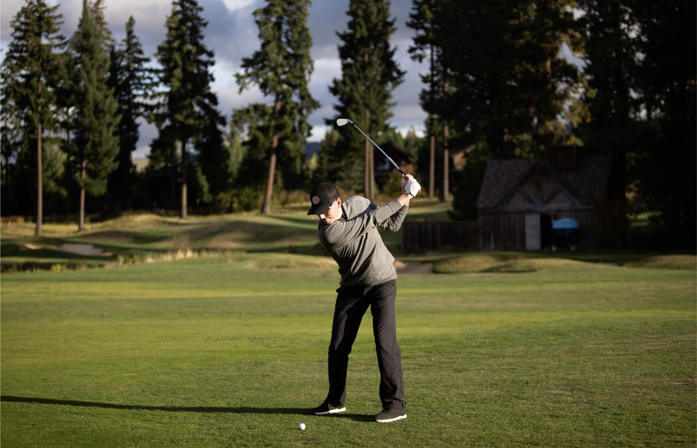 A person in a gray sweater and black pants swings a golf club on a grassy course at Suncadia Resort in Washington, with trees and a small building in the background.