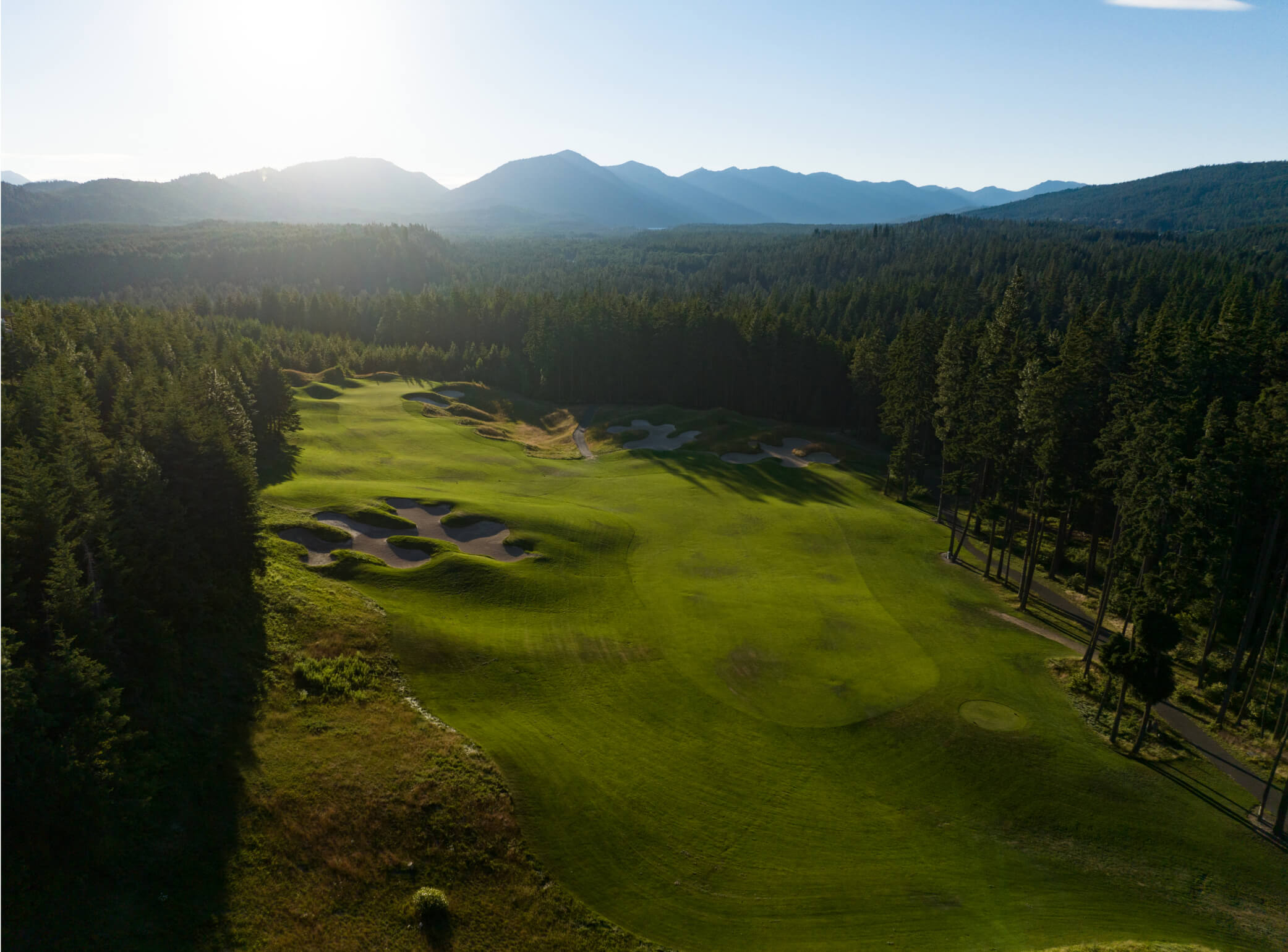 Aerial view of Suncadia's golf course with sand bunkers and lush greens, surrounded by dense forest and majestic mountains under a clear sky.