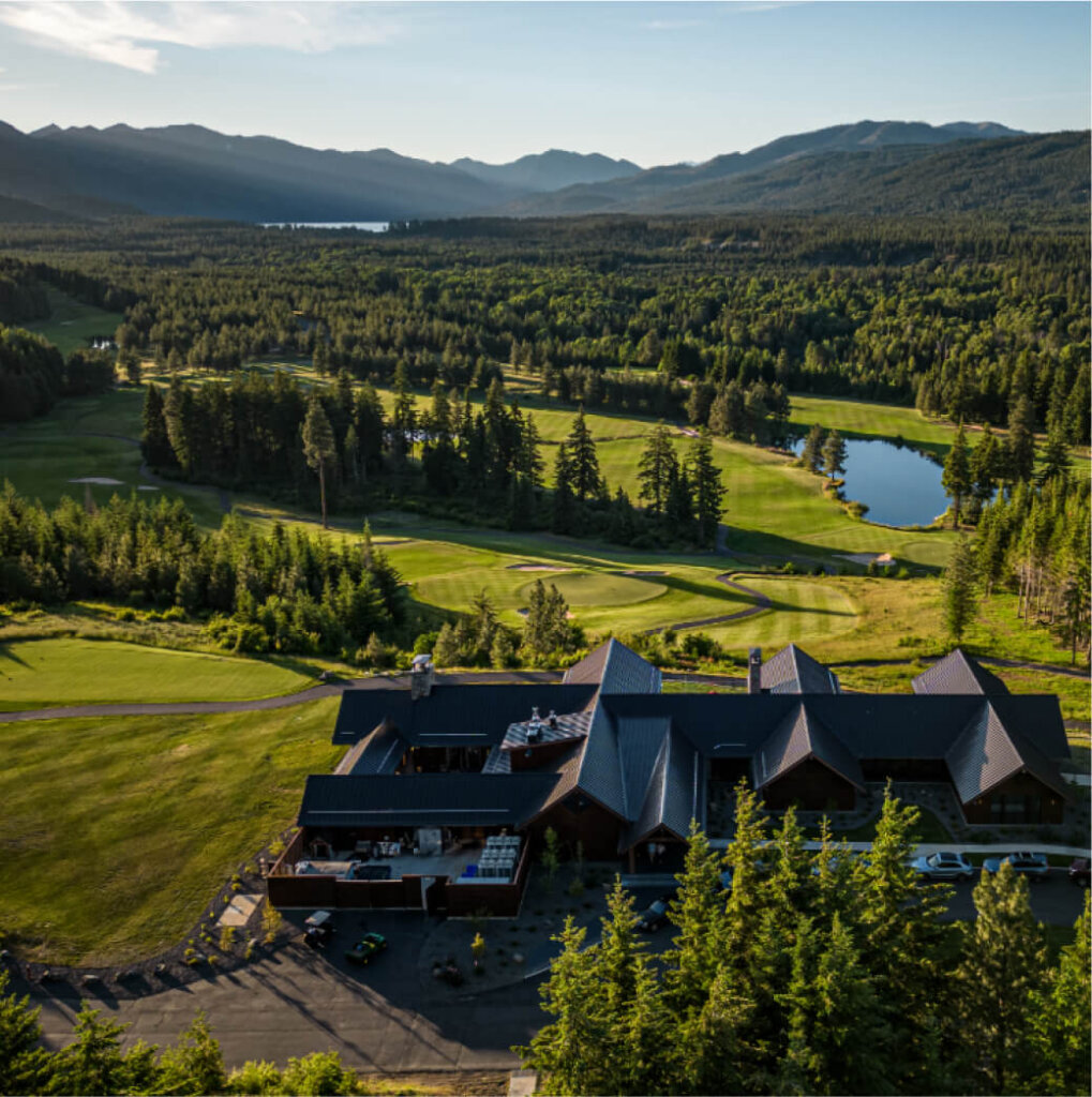 Aerial view of a large building surrounded by a pristine golf course, lush trees, and a small lake, set against the backdrop of majestic mountains on a sunny day. Discover the beauty of Suncadia real estate in this stunning landscape.