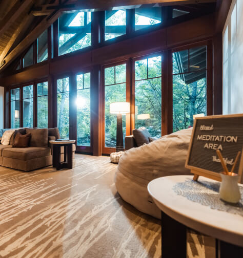 A cozy meditation area with a bean bag, sofa, and table with sign. Large windows showcase a forest view reminiscent of Suncadia homes, and there's a floor lamp for lighting.