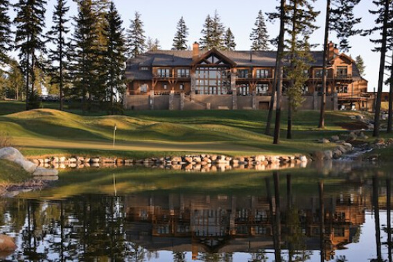 A large wooden lodge is reflected in a pond, nestled within the serene beauty of Suncadia Resort Washington, surrounded by tall trees and a grassy lawn.