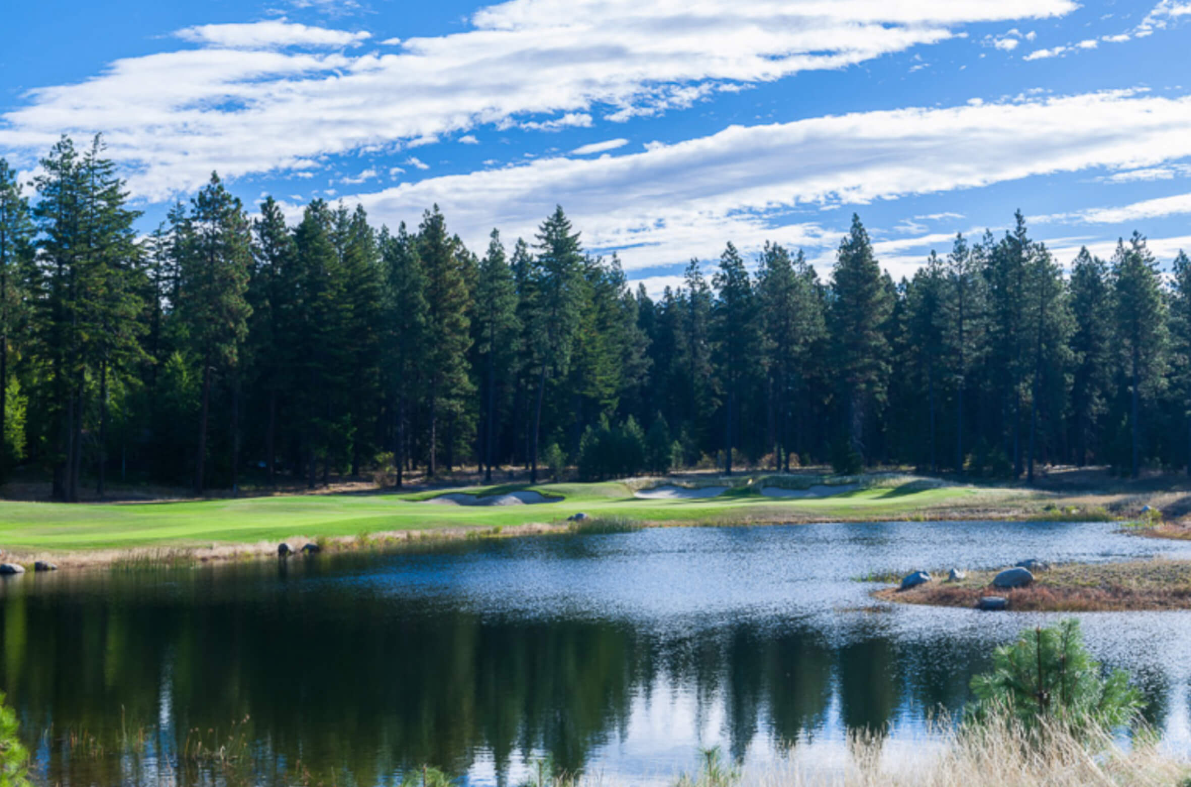 A serene golf course with a pond in the foreground, surrounded by tall evergreen trees under a partly cloudy blue sky, offers a picturesque setting at Suncadia Resort. Enjoy nearby Cle Elum restaurants and explore luxurious Suncadia homes for an unforgettable experience.