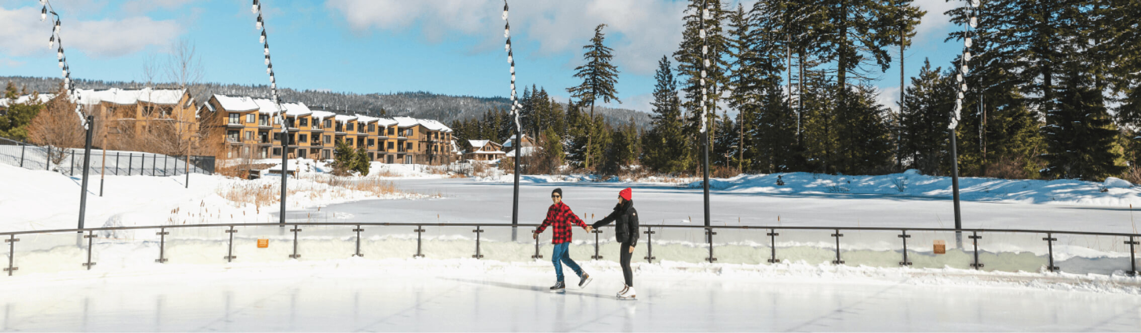 Two people ice skating on an outdoor rink surrounded by snow and pine trees, with the charming backdrop of Suncadia Resort Washington.