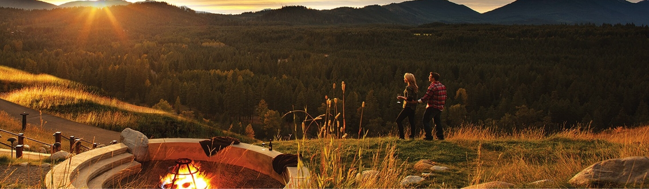 Two people stroll near a fire pit on a grassy hill at sunset, with the serene backdrop of Suncadia real estate nestled among forest and mountains.