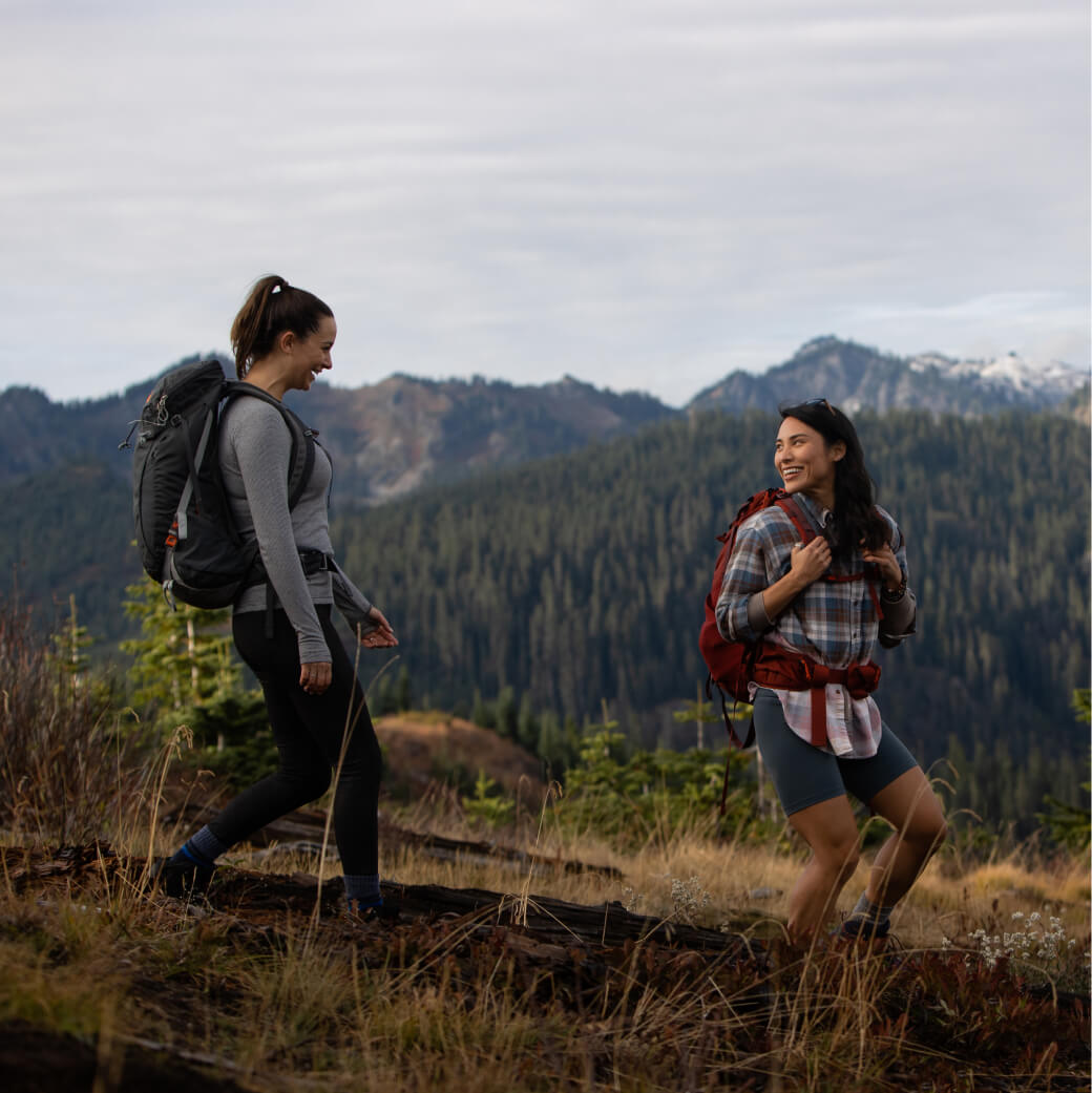 Two people hiking on a trail in a mountainous area near Suncadia Resort, clad in backpacks and outdoor gear. Forested hills embrace the path under a cloudy sky, as they dream of unwinding at Cle Elum restaurants after their adventure.