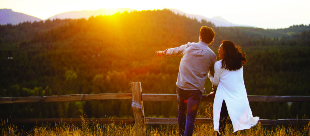 A couple stands by a wooden fence, pointing towards the sunset over a forested mountain landscape, dreaming of suncadia real estate where nature's beauty meets luxurious living.