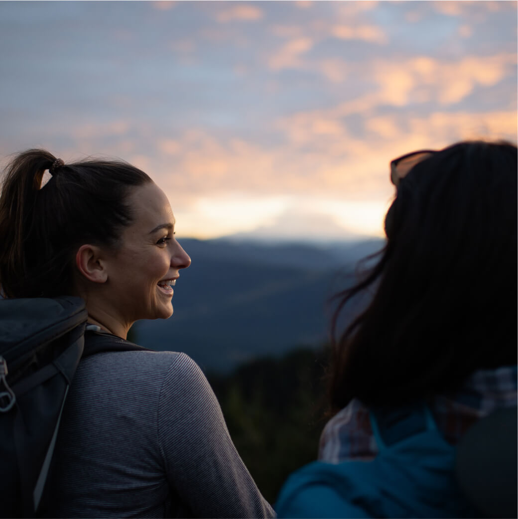 Two people with backpacks enjoy a scenic sunset view over the distant mountains, one smiling at the other, as they reminisce about their day exploring Suncadia Resort Washington.