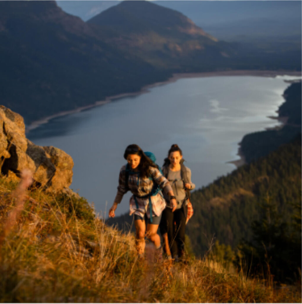 Three people hike up a grassy hill at Suncadia Resort, overlooking a large lake framed by majestic mountains as the sunset casts a golden glow.