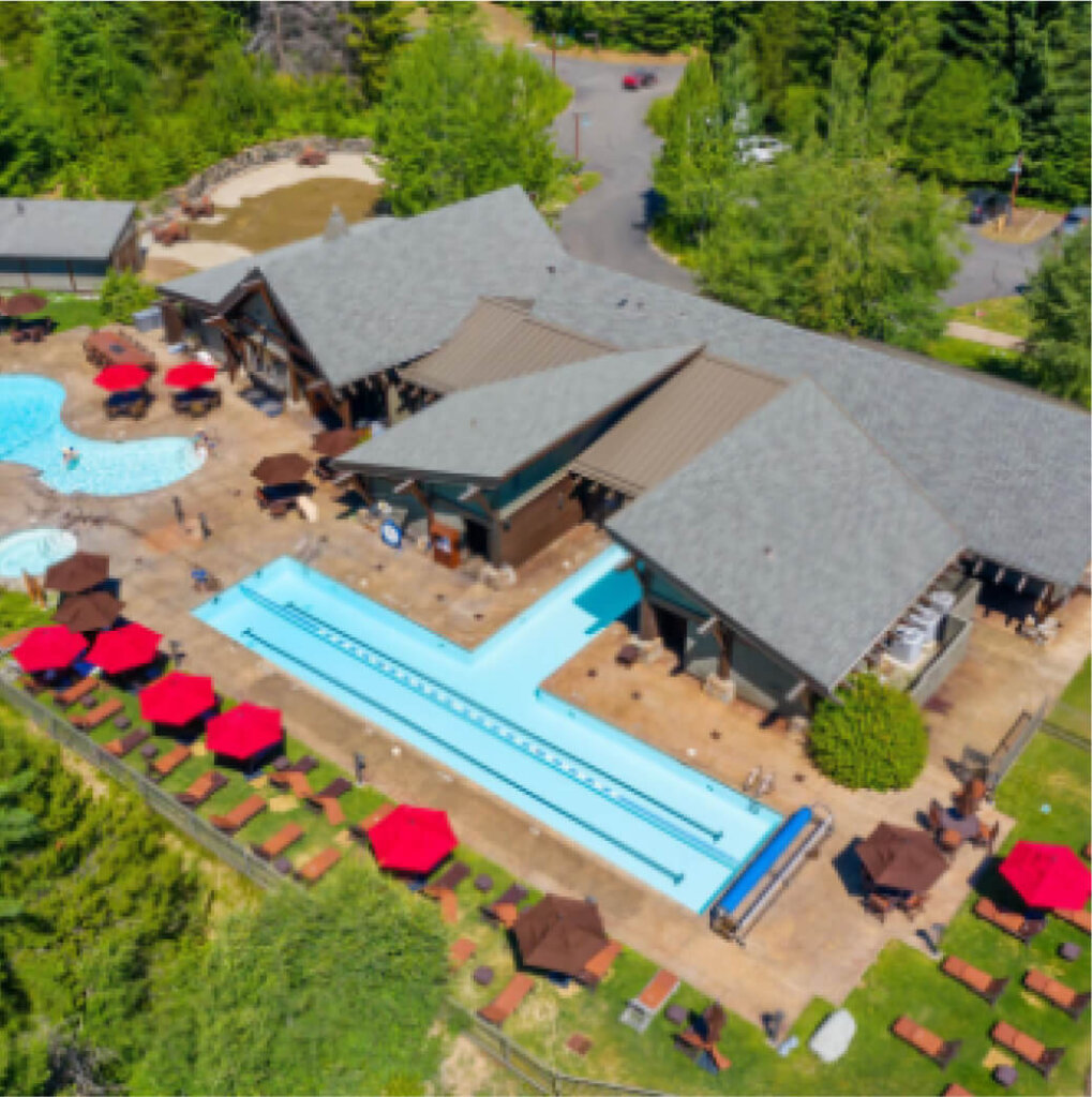 Aerial view of a Suncadia lodge with a T-shaped pool, surrounded by lounge chairs, vibrant red umbrellas, and lush greenery.