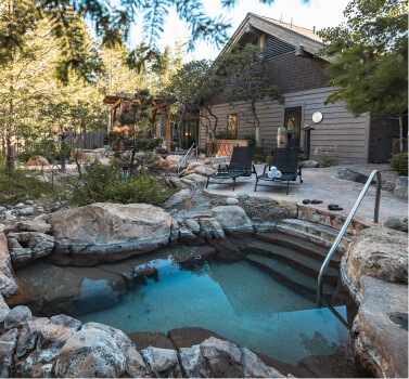 A charming outdoor hot tub nestled among rocks and trees, set against a rustic cabin backdrop. Nearby, two lounge chairs await beside a pathway. This tranquil scene is reminiscent of the serene lifestyle found in Suncadia homes and the vibrant offerings of Cle Elum restaurants.