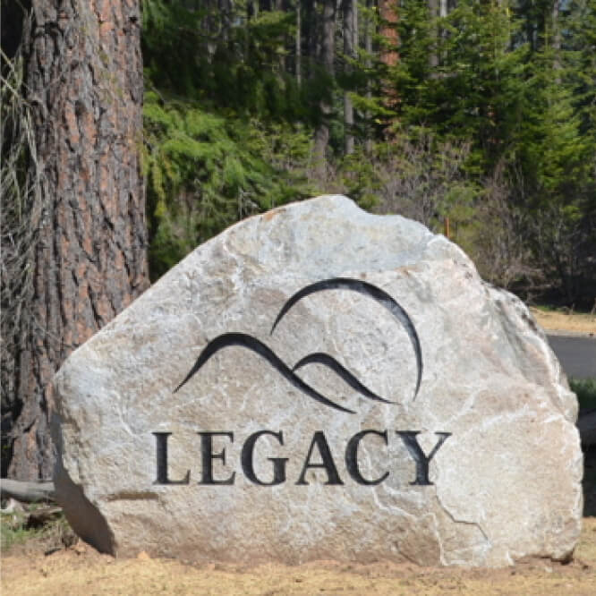 A stone sign with the word "LEGACY" and a stylized wave design engraved on it stands proudly amidst the lush greenery, capturing the essence of Suncadia's beautiful landscape, where exquisite real estate meets the charm of nearby Cle Elum restaurants.