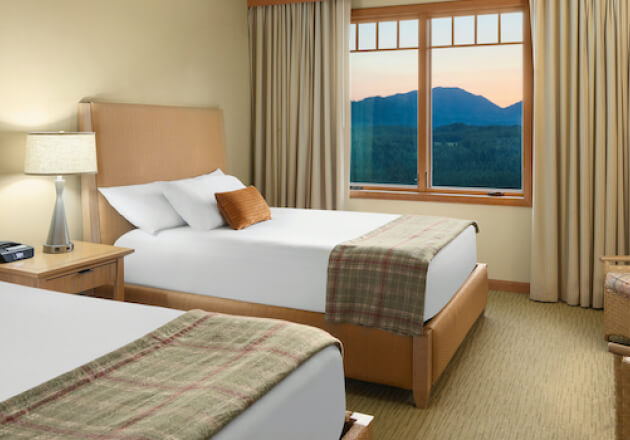 The hotel room features two cozy beds, a nightstand, and a lamp, all perfectly positioned near a window that frames the stunning mountain sunset, reminiscent of Suncadia Resort's breathtaking vistas.