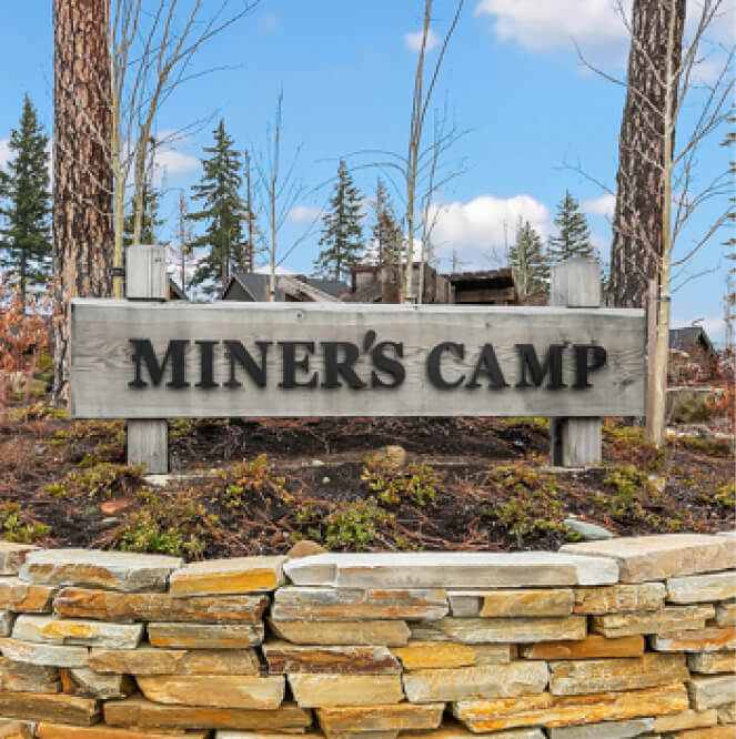 A wooden sign reading "MINER'S CAMP" is mounted on a stone wall, with the picturesque backdrop of trees and houses, evoking the charm of Suncadia Resort, Washington. Perfect for those exploring Suncadia real estate or dreaming of owning a home in this serene landscape.