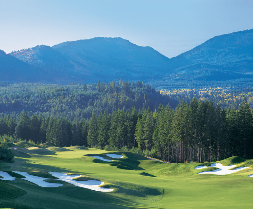 A lush green golf course with sand bunkers sits within the enchanting Suncadia landscape, framed by forested hills and mountains under a clear blue sky.