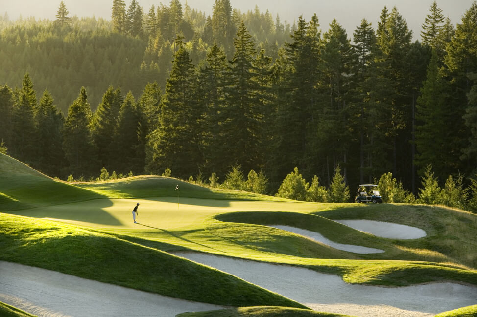 A golfer at the Suncadia Resort's sunlit course prepares to putt, surrounded by sand traps and lush green trees in the background.