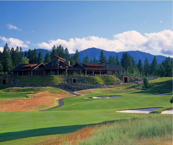 A golf course with a large clubhouse at Suncadia Resort, Washington, surrounded by trees and mountains under a partly cloudy sky. Just minutes from Cle Elum restaurants and charming Suncadia homes, it's the perfect blend of leisure and natural beauty.