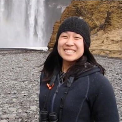 A person smiles while standing in front of a large waterfall and rocky terrain, wearing a black beanie and a dark jacket with binoculars around their neck, perhaps scouting the breathtaking views of Suncadia Resort in Washington after exploring nearby Cle Elum restaurants.