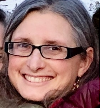 A woman with glasses smiles warmly, wearing a red jacket that complements her gray and brown hair. Her radiant expression seems to capture the serene allure of Suncadia Resort, Washington, as if she's just returned from exploring potential Suncadia homes under the pristine skies.