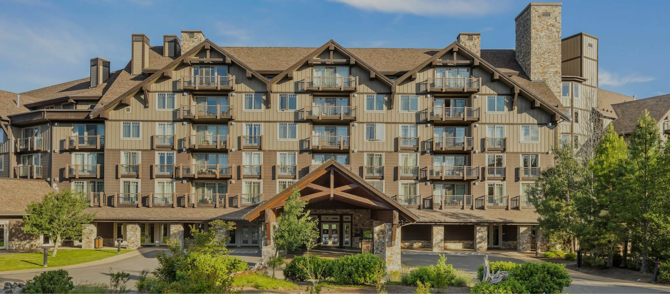 A large multi-story lodge at Suncadia Resort features a wooden entrance, multiple balconies, and stone accents, all surrounded by lush greenery.