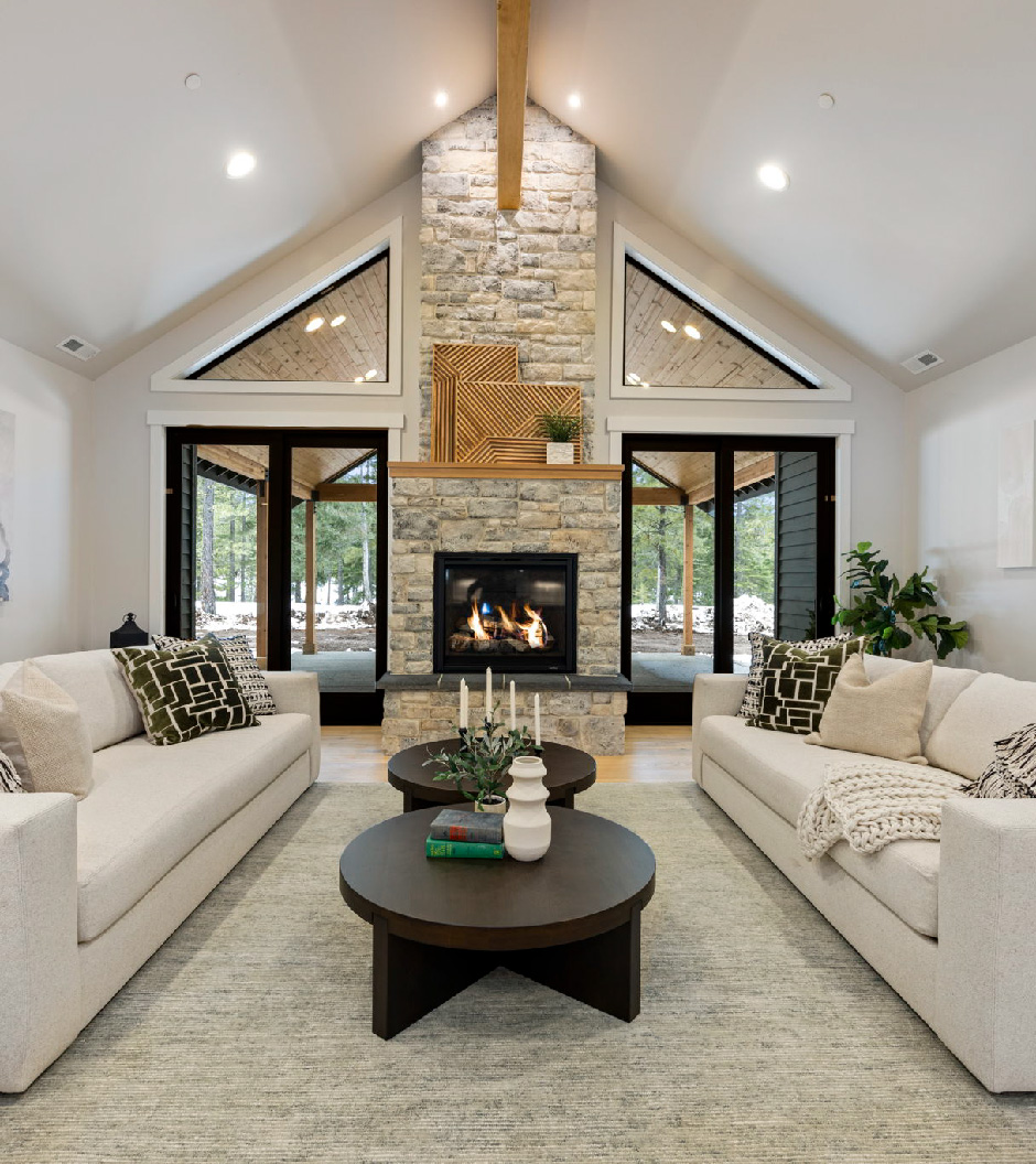 A cozy living room with a stone fireplace, two white sofas, and round coffee tables showcases the charm of Suncadia homes. Large windows reveal a wooded view outside, echoing the serene beauty of Suncadia Resort.