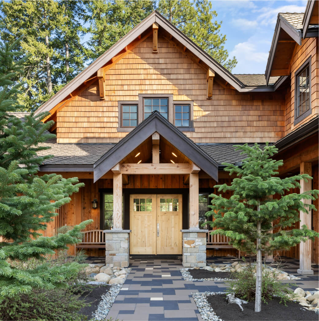 A charming wooden house with a gabled roof, stone columns, and a covered entrance is nestled among trees and a patterned walkway. It embodies the rustic elegance of Suncadia homes in the breathtaking landscape of Suncadia Resort Washington.