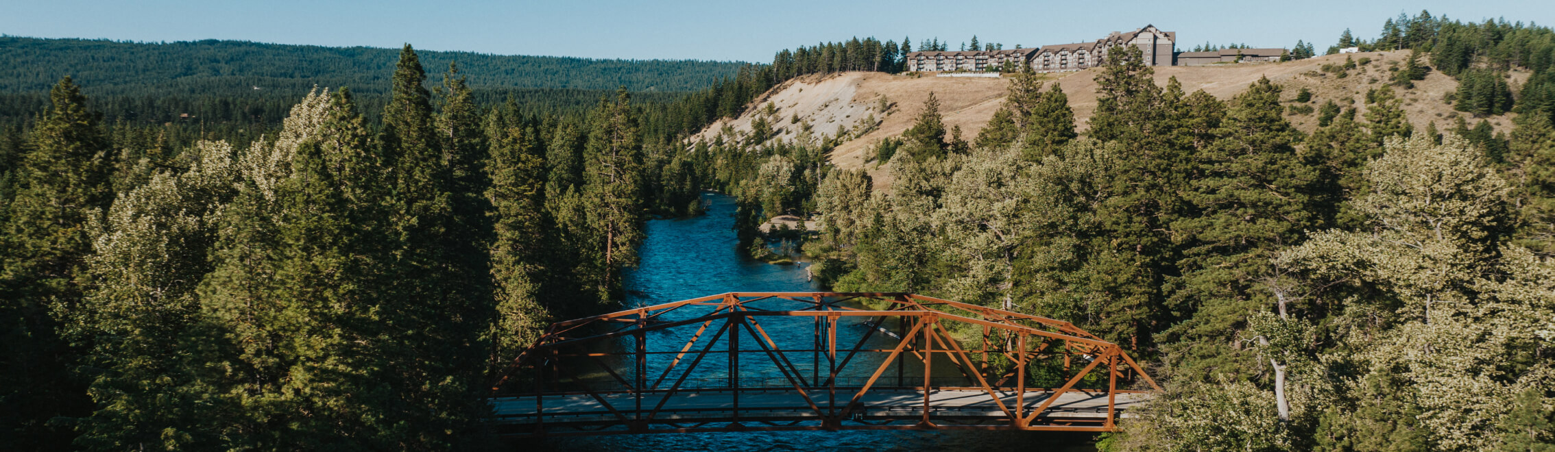 A rust-colored bridge spans a river surrounded by dense pine forests, with a large building atop a hill in the background, reminiscent of the serene beauty found near Suncadia Resort Washington.