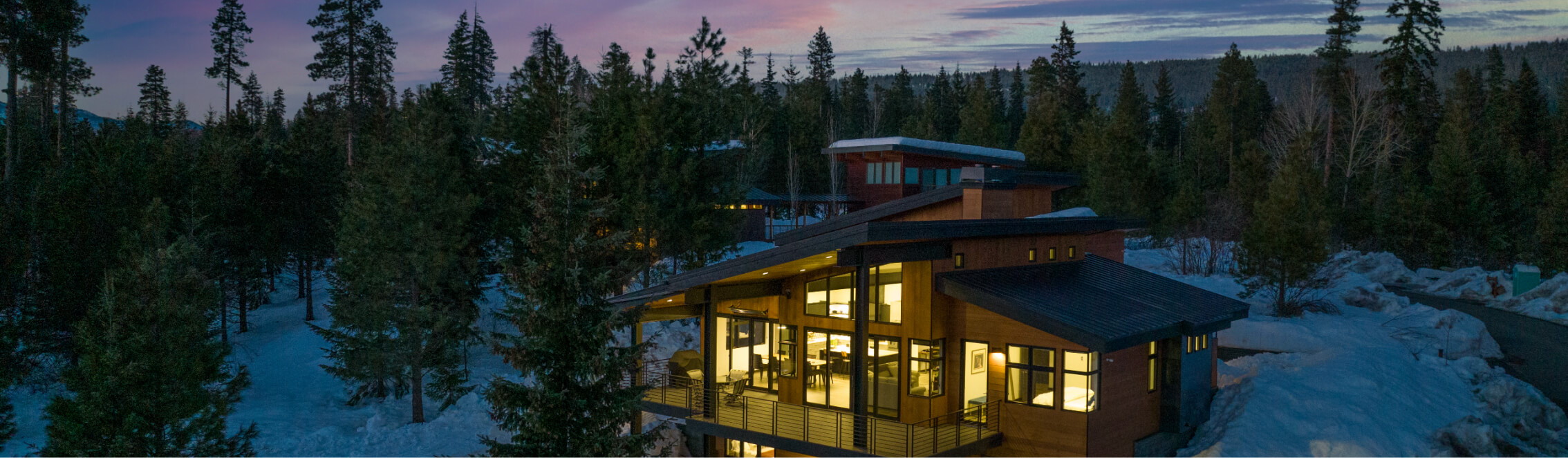 A modern wooden house with large windows is nestled among snow-covered trees at dusk in the tranquil Suncadia resort, Washington, set against a stunning purple and blue sky.