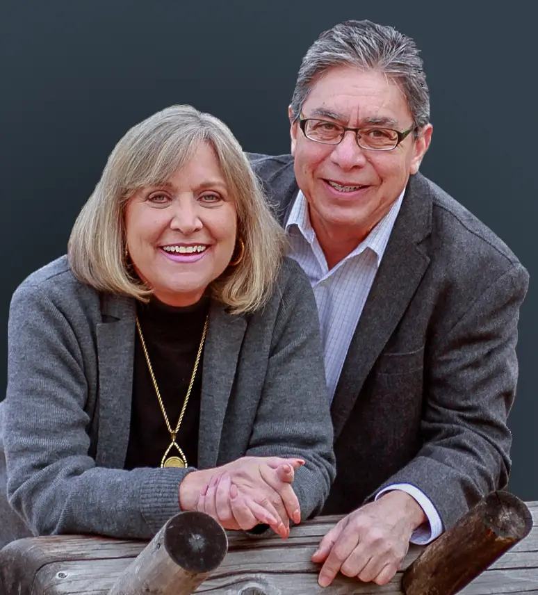 A smiling man and woman in gray jackets lean on wooden railings against a dark background, capturing the serene ambiance of Suncadia Resort.