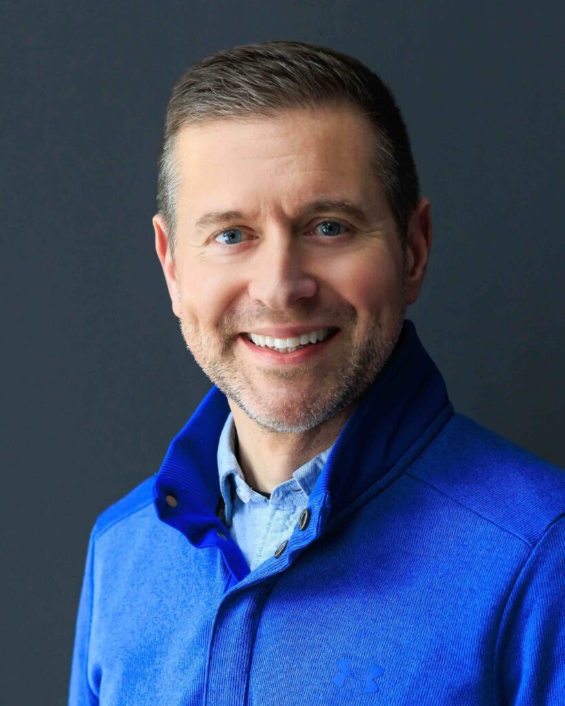 A person wearing a blue jacket smiles against a dark gray background, reminiscent of a cozy evening at Suncadia Resort, Washington.