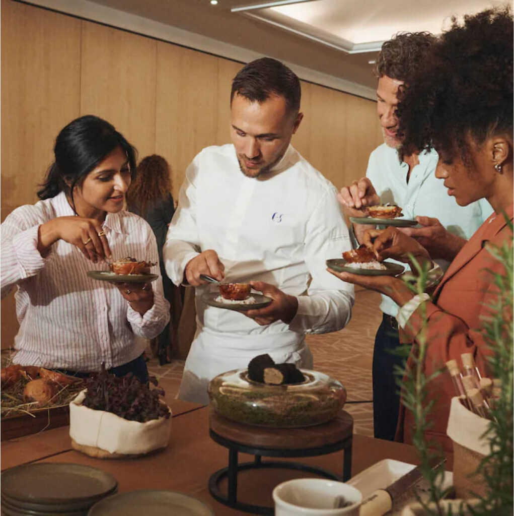 A group of four people stand together, savoring food from small plates at an indoor event in Suncadia. Nearby, a table with various dishes beckons more attendees to explore the local flavors and enjoy the community spirit that Suncadia homes are known for fostering.