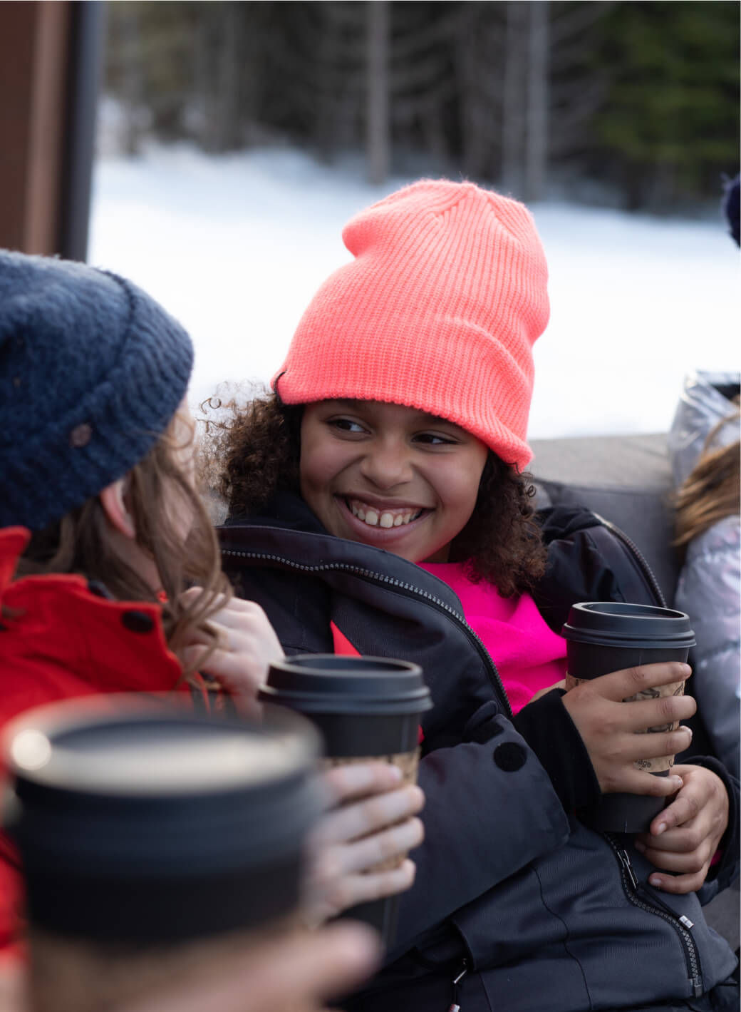 Children in winter clothing, holding insulated cups, smile and chat outdoors with a snowy backdrop at Suncadia Resort.