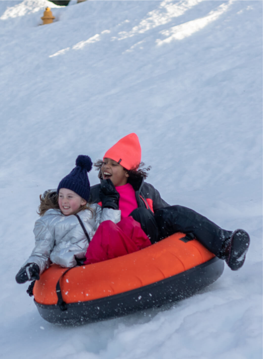 Two people in winter clothing are joyfully sledding on an orange inflatable tube down a snowy hill, enjoying the stunning views near Suncadia Resort.