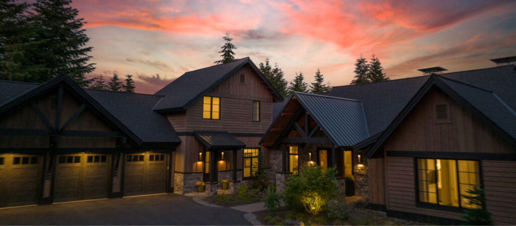 A rustic wooden house with a steep roof and multiple gables, illuminated by outdoor lights, set against a vibrant sunset sky and surrounded by tall trees. This serene escape embodies the charm of Suncadia real estate in Washington, offering a perfect blend of nature and comfort.