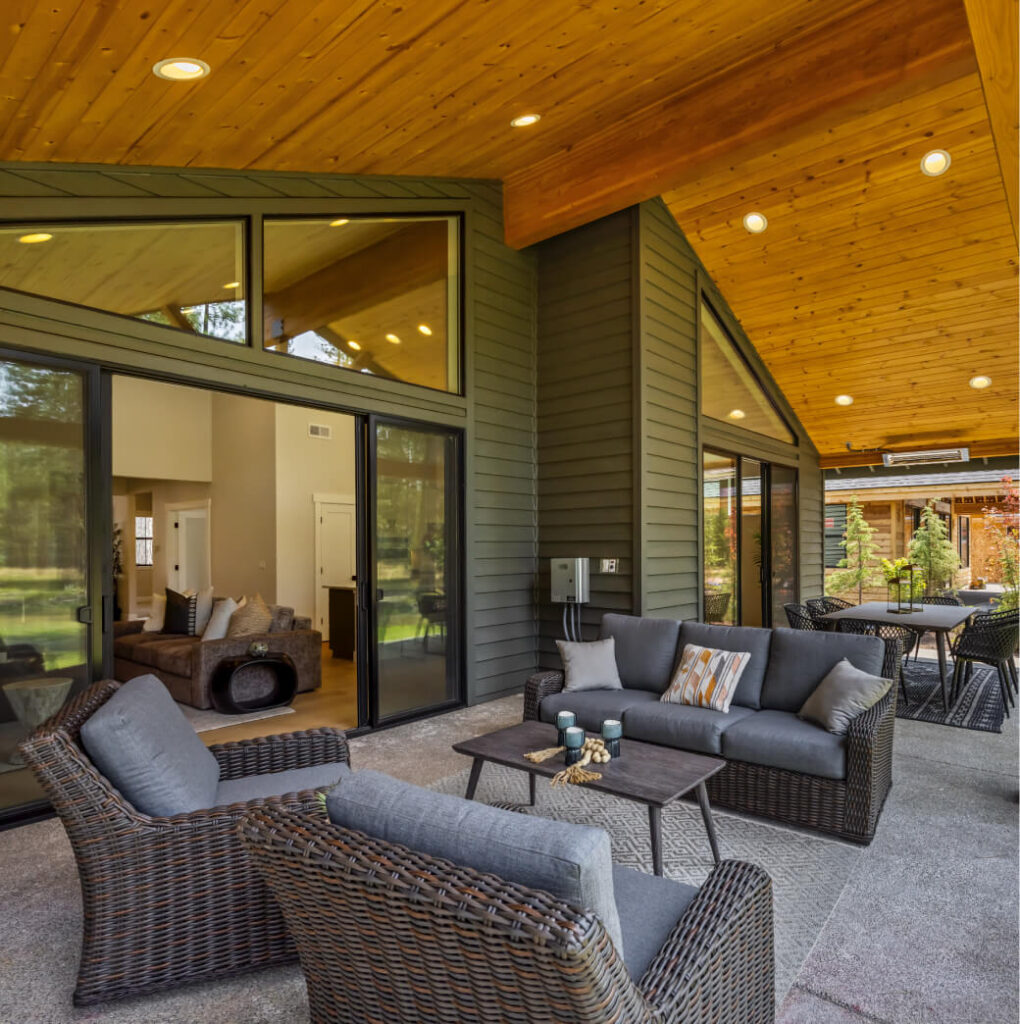 Experience a serene atmosphere with this covered patio featuring gray wicker furniture and plush cushions. The wooden ceiling, adorned with recessed lights, adds elegance. Large sliding glass doors lead to an inviting indoor living area, reminiscent of the luxurious style of Suncadia Resort living.