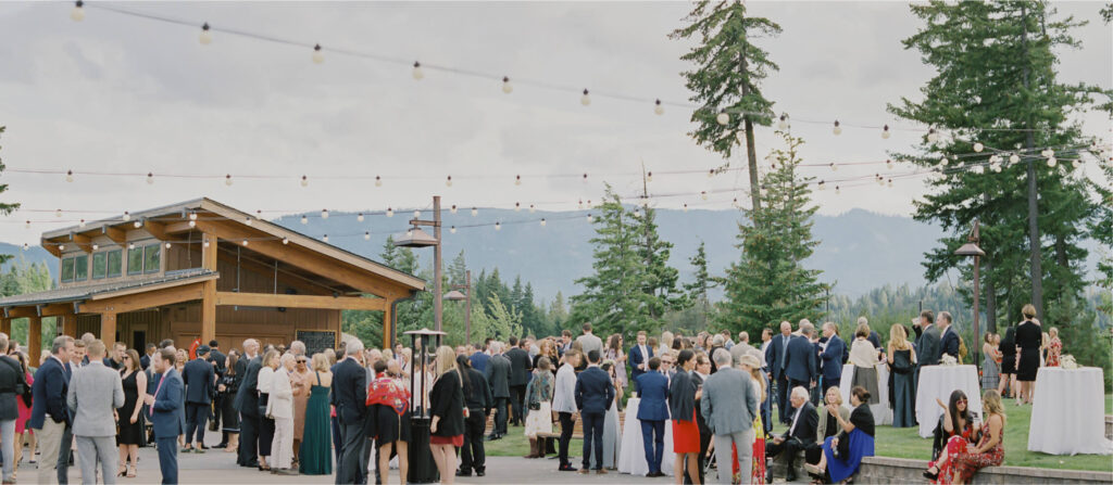 An outdoor gathering at Suncadia Resort, Washington, features many people dressed in formal attire beneath string lights overhead, surrounded by the breathtaking scenery of trees and mountains.