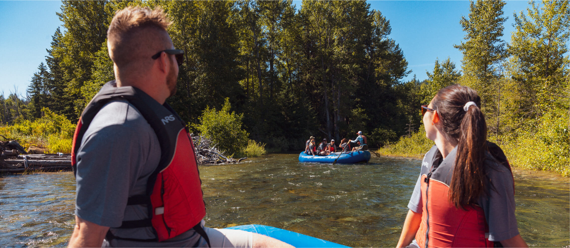 Two people in life vests sit in a raft on a river near Suncadia, watching another raft with several passengers navigate through the water, surrounded by trees. After their adventure, they plan to visit Cle Elum restaurants for a delicious meal.