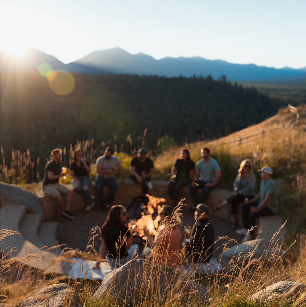 A group of people sitting around a fire pit outdoors, basking in the glow of sunset against the majestic mountain backdrop near Suncadia Resort.