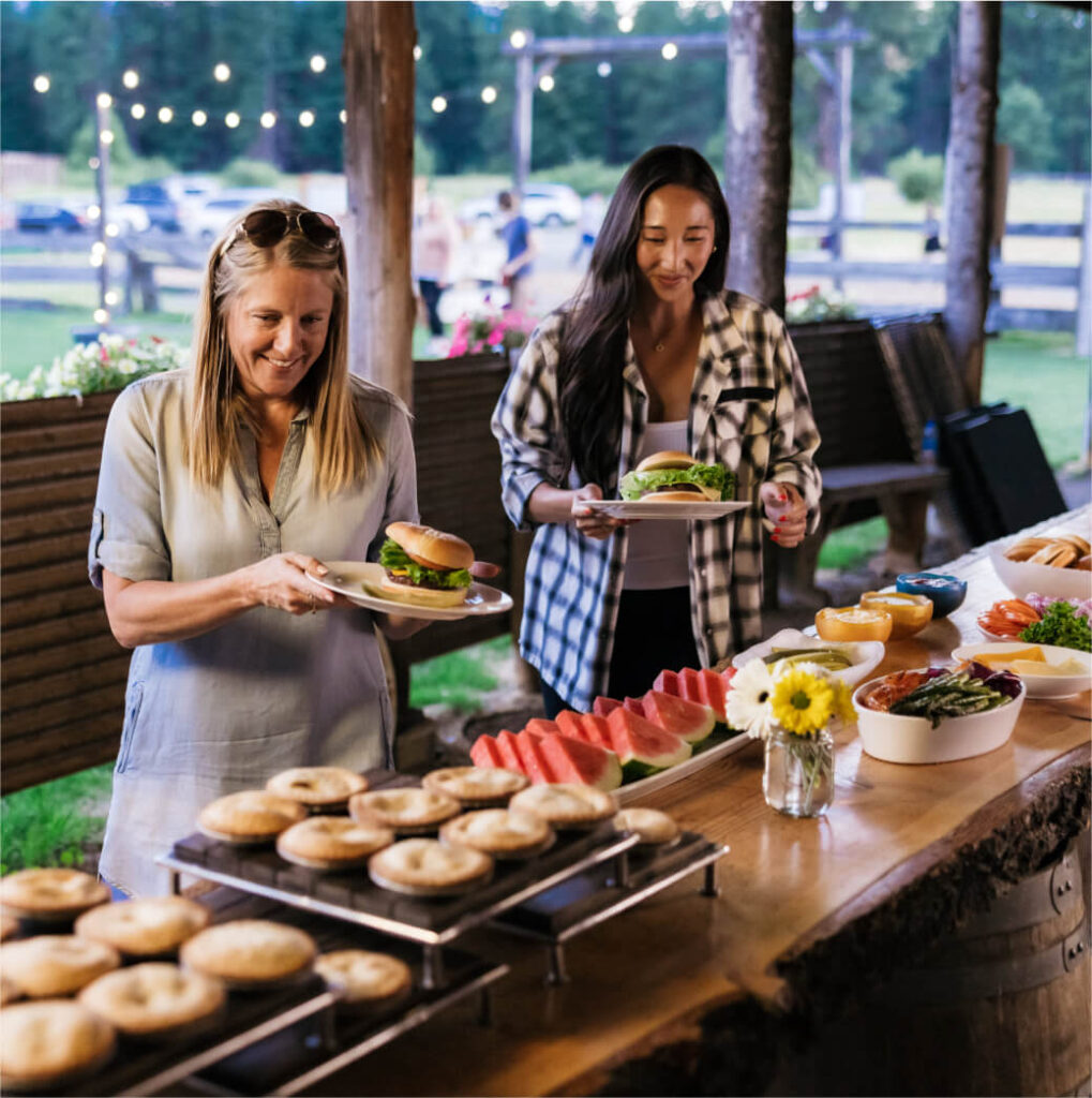 Two people stand at a buffet table outdoors, selecting from an array of food items with bagels and watermelon prominently displayed. In the backdrop of this idyllic Suncadia Resort setting, string lights twinkle among the trees, creating a warm and inviting atmosphere.