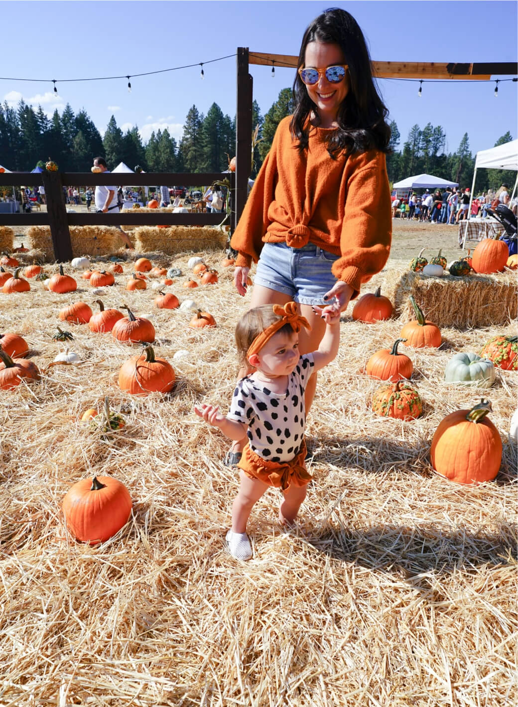 A woman and a child stroll through a pumpkin patch at Suncadia Resort Washington, surrounded by hay and pumpkins. The woman wears sunglasses and an orange sweater; the child sports a bow and a polka dot outfit, perfectly capturing the essence of autumn in this charming setting.