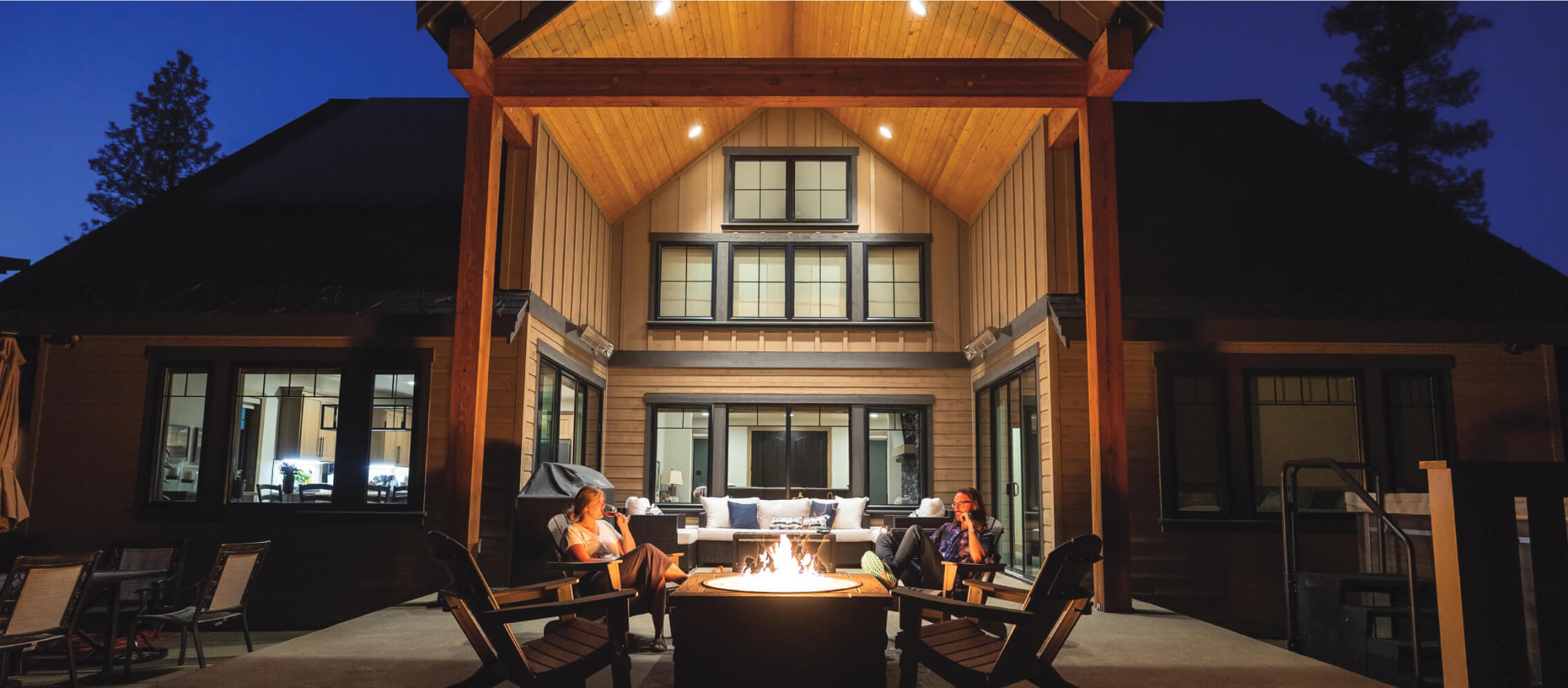 Two people sit around a fire pit on a patio at night, framed by a wooden structure, reminiscent of cozy evenings in Suncadia homes. The house is lit in the background.