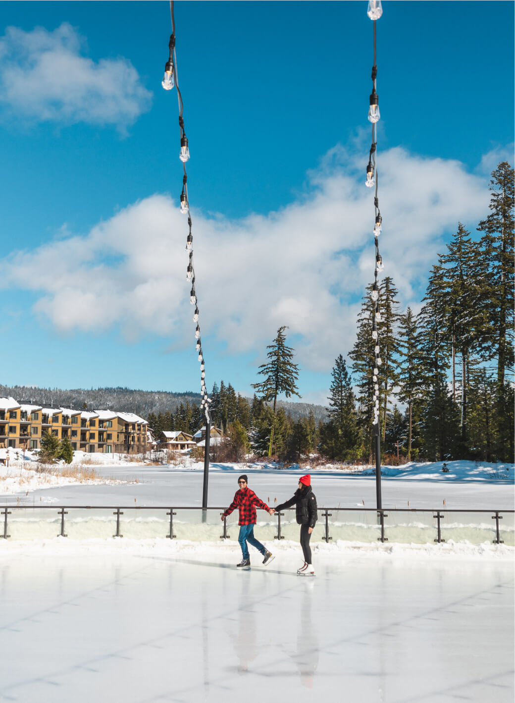 Two people ice skating on an outdoor rink at Suncadia Resort, surrounded by trees and charming buildings under a clear blue sky, with string lights twinkling overhead.