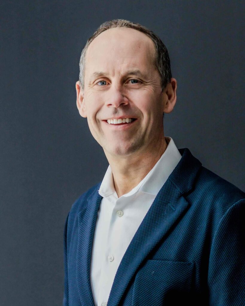 Against a dark gray backdrop, a person in a blue suit and white shirt beams with a smile, perhaps reflecting the charm of Cle Elum restaurants.