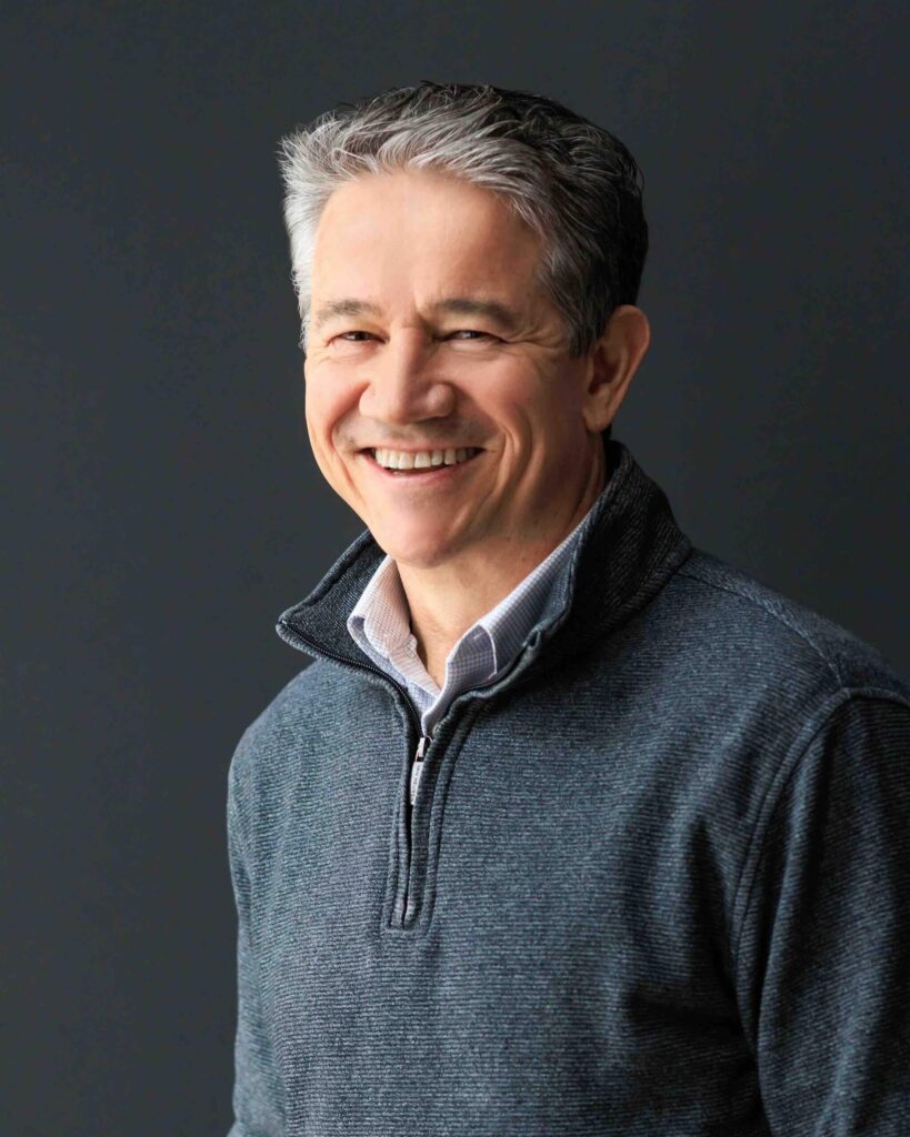 A man with short gray hair is smiling, wearing a dark gray zip-up sweater over a light collared shirt. The background is a plain dark color, reminiscent of the tranquil ambiance often found at Suncadia Resort.