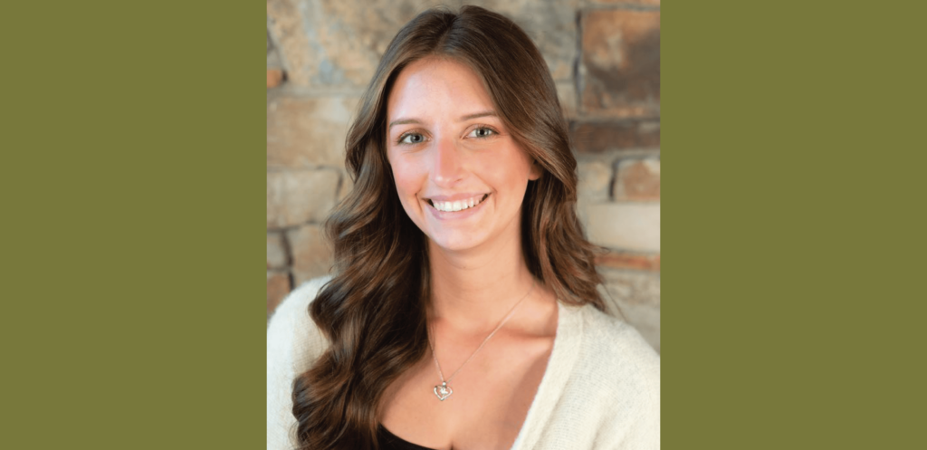 A young woman with long brown hair smiles at the camera. She is wearing a white top and a necklace, standing against a stone wall at Suncadia Resort.