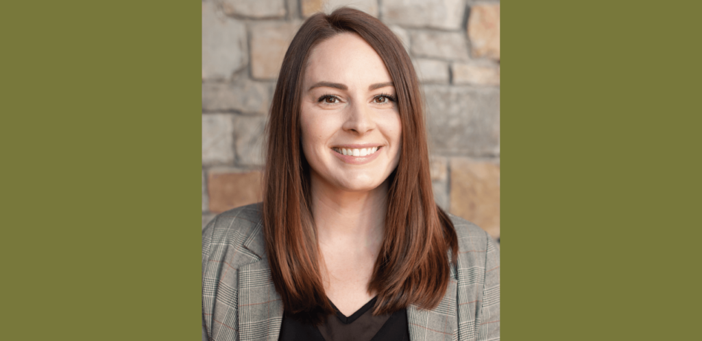 A person with long brown hair, wearing a plaid blazer and black top, smiles in front of a stone wall, reminiscent of the cozy charm found in Suncadia homes.