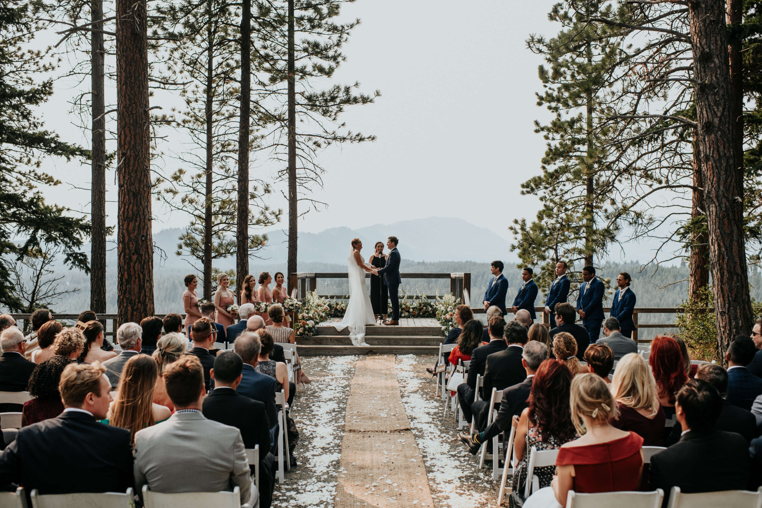 A wedding ceremony unfolds outdoors at Suncadia Resort, with a couple standing at the altar. Guests are seated on both sides, surrounded by tall trees and distant mountains, creating a perfect backdrop for the union.